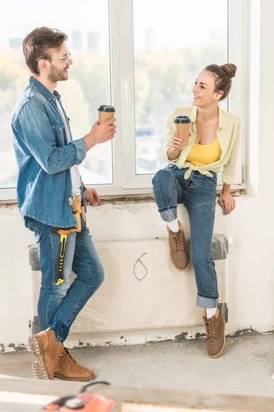 Feliz jovem casal segurando copos de papel com café para ir e sorrindo uns aos outros em nova casa — Fotografia de Stock