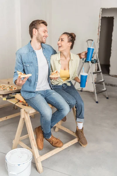 Feliz pareja joven con pizza y vasos de papel sentados juntos y sonriendo entre sí durante la reparación - foto de stock