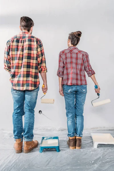 Vue arrière du jeune couple tenant des rouleaux de peinture et regardant le mur dans une nouvelle maison — Photo de stock