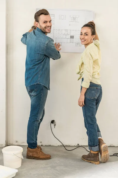 Glückliches junges Paar mit Blaupause an der Wand und lächelnd in die Kamera im neuen Haus — Stockfoto