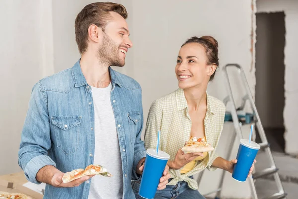 Felice giovane coppia in possesso di bicchieri di carta con cannucce e fette di pizza nel nuovo appartamento — Foto stock