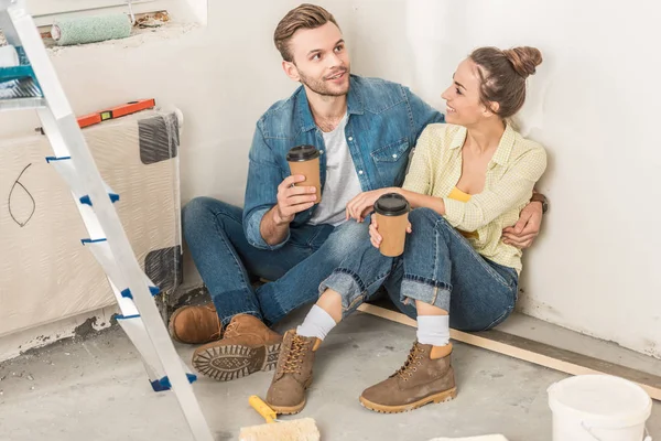 Blick aus der Vogelperspektive auf ein lächelndes junges Paar, das Pappbecher hält und in einer neuen Wohnung auf dem Boden sitzt — Stockfoto