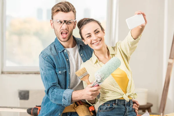 Feliz joven pareja sosteniendo herramientas y tomando selfie con teléfono inteligente durante la renovación - foto de stock