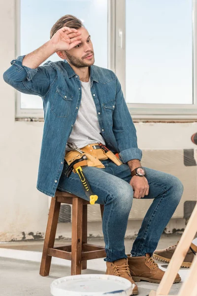 Stanco giovane uomo con cintura degli attrezzi seduto sulla sedia durante la riparazione della casa — Foto stock