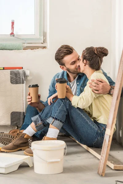 Young couple holding paper cups and kissing while sitting on floor in new house — Stock Photo