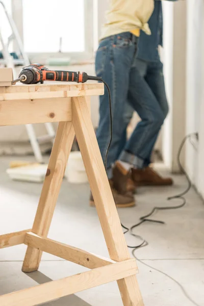 Taladro eléctrico en mesa de madera y pareja joven de pie cerca de la pared detrás - foto de stock