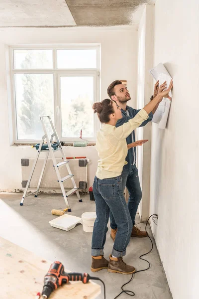 Vista lateral de la pareja joven sosteniendo el plano en la pared durante la renovación - foto de stock