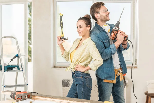 Feliz joven pareja sosteniendo herramientas y de pie espalda con espalda durante la renovación - foto de stock