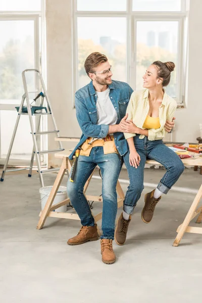 Feliz joven pareja abrazándose y sonriendo entre sí mientras se sienta en la mesa con herramientas - foto de stock