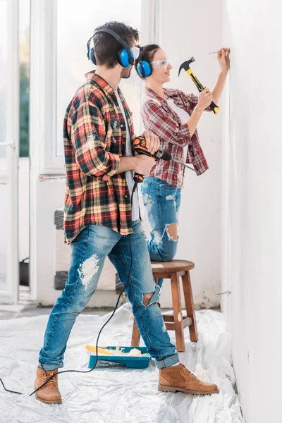 Вид сбоку молодого человека, держащего электрическую капельницу и смотрящего на улыбающуюся жену, забивающую гвоздь в новой квартире — стоковое фото