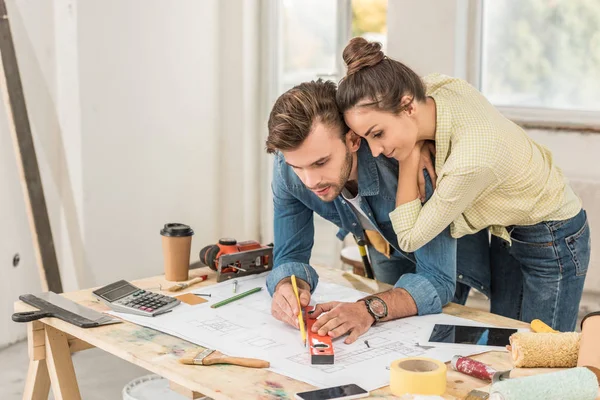 Junge Frau schaut Ehemann beim Markieren von Bauplänen mit Niveauwerkzeug in neuem Haus an — Stockfoto