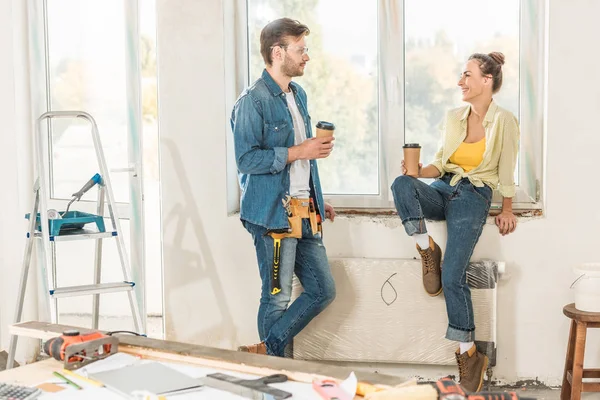 Feliz joven pareja sosteniendo café para ir y sonriendo el uno al otro durante la reparación - foto de stock