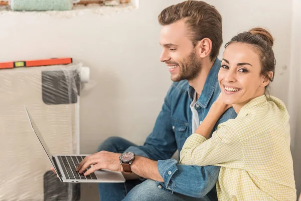 Щасливий молодий чоловік використовує ноутбук і красива дружина посміхається на камеру в новому будинку — стокове фото