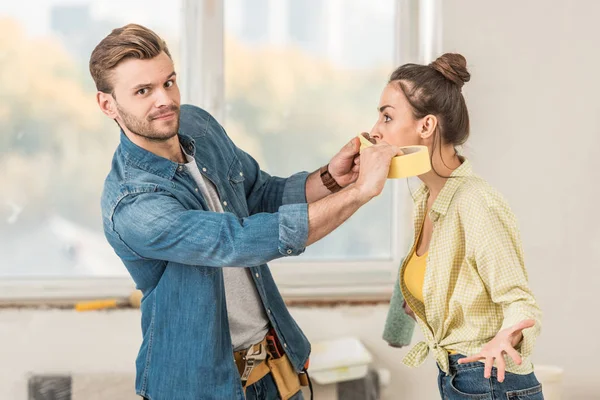 Man holding adhesive tape near face of his wife and looking at camera during home repair — Stock Photo