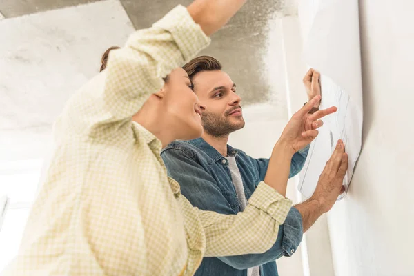 Vista de ángulo bajo de la joven pareja sonriente sosteniendo el plano en la pared durante la reparación — Stock Photo