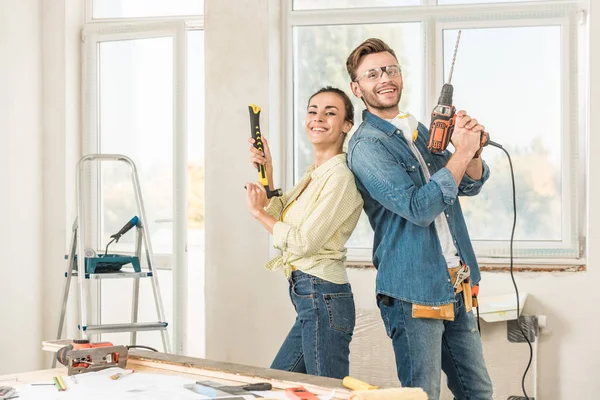 Feliz joven pareja sosteniendo herramientas y sonriendo a la cámara durante la reparación de la casa - foto de stock