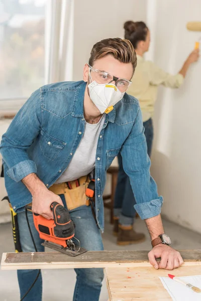 Young man in protective workwear using electric jigsaw and looking at camera — Stock Photo