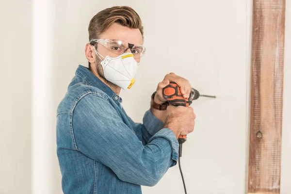 Jeune homme en vêtements de travail de protection en utilisant une perceuse électrique et en regardant la caméra pendant la réparation de la maison — Photo de stock