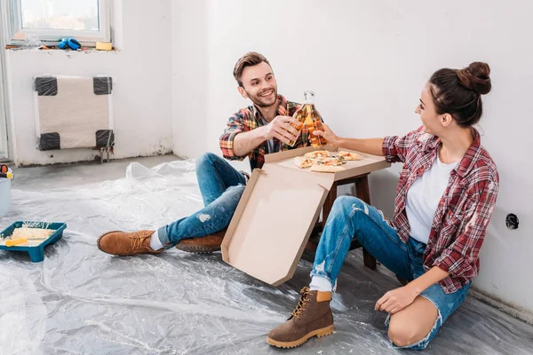 Счастливая молодая пара звон бутылки пива и есть пиццу, сидя на полу в новой квартире — стоковое фото