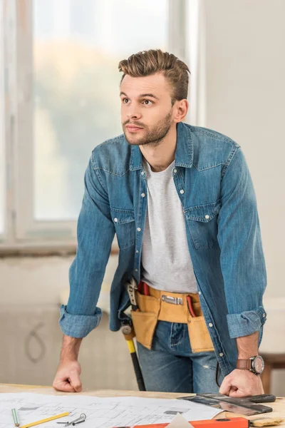 Beau jeune homme avec ceinture d'outils penché à la table et regardant loin à la nouvelle maison — Photo de stock