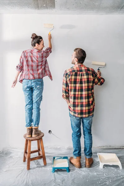 Vue arrière du mur de peinture heureux jeune couple avec des rouleaux de peinture — Photo de stock