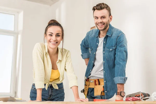 Feliz joven pareja inclinada a la mesa con herramientas y sonriendo a la cámara durante la reparación de la casa — Stock Photo