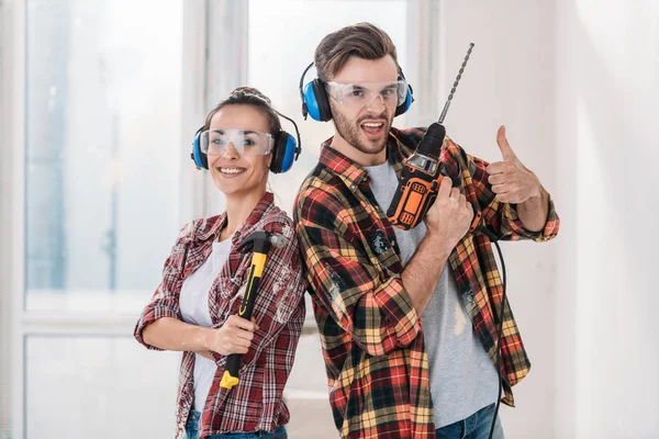 Glückliches junges Paar in karierten Hemden, Werkzeug in der Hand und in die Kamera blickend — Stockfoto