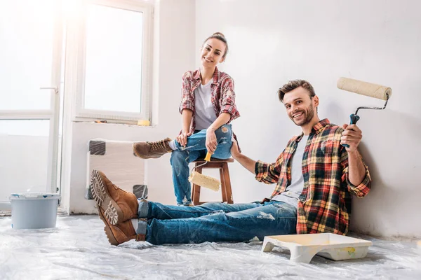 Heureux jeune couple tenant des rouleaux de peinture et souriant à la caméra pendant la rénovation — Photo de stock