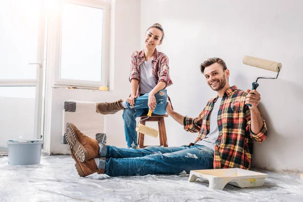 Feliz joven pareja sosteniendo rodillos de pintura y sonriendo a la cámara durante la renovación - foto de stock