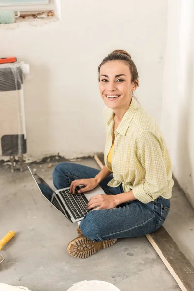 Приваблива молода жінка, використовуючи ноутбук і посміхатися камери під час облаштування будинку — стокове фото