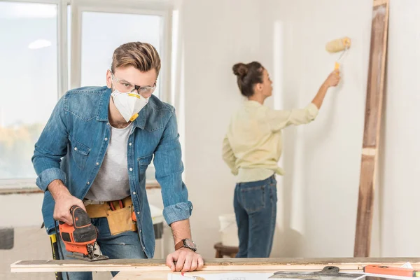 Young man in protective workwear using electric jigsaw and looking at camera while wife painting wall behind — Stock Photo