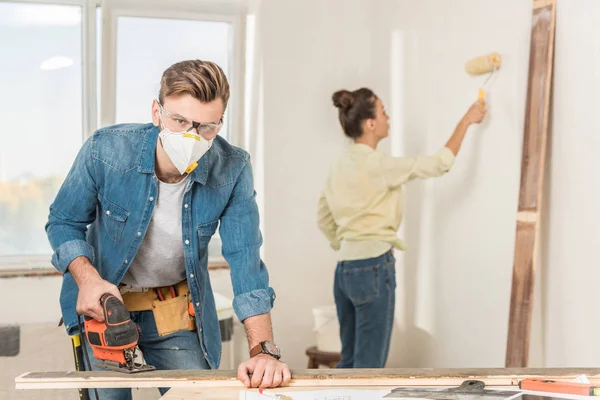 Joven en ropa de trabajo protectora usando rompecabezas eléctrico y mirando a la cámara mientras la esposa pintando la pared detrás - foto de stock