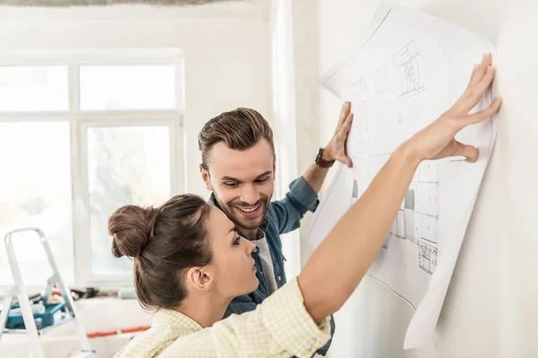 Jeune couple tenant le plan au mur lors de la rénovation dans un nouvel appartement — Photo de stock