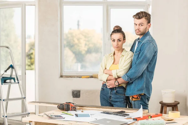 Seria pareja joven abrazando y mirando a la cámara durante la reparación de la casa - foto de stock