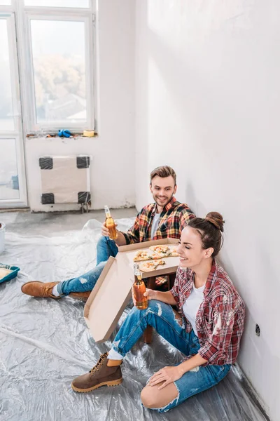 Високий кут зору щасливої молодої пари п'є пиво і їсть піцу під час благоустрою будинку — стокове фото
