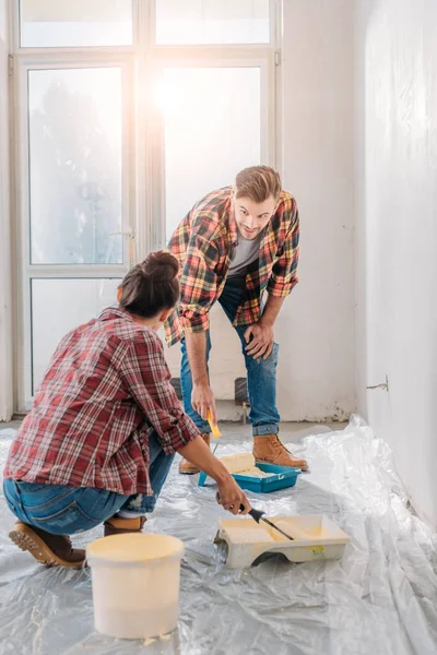 Jeune couple en chemises à carreaux tenant des rouleaux de peinture et mur de peinture — Photo de stock