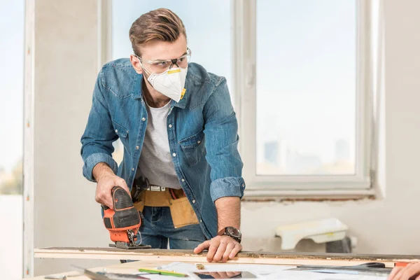 Young man in protective workwear using electric jigsaw while making repair at home — Stock Photo