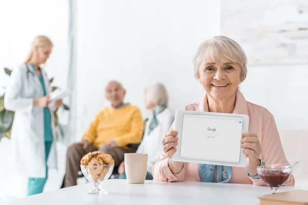 Senior femme tenant tablette numérique avec google app dans la maison de soins infirmiers avec des gens à l'arrière-plan — Photo de stock