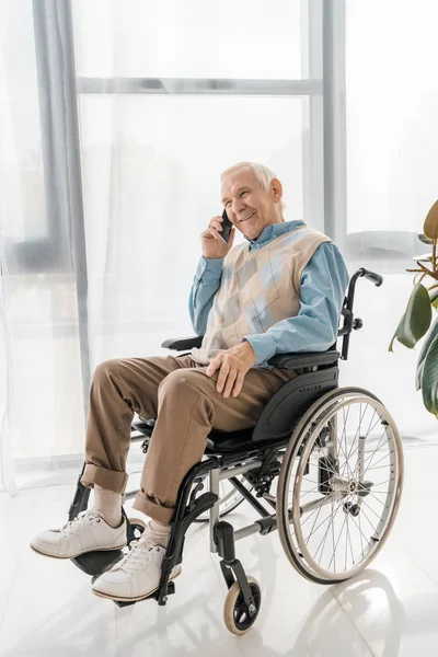 Старший улыбающийся мужчина сидит в инвалидной коляске и разговаривает по смартфону в доме престарелых — стоковое фото