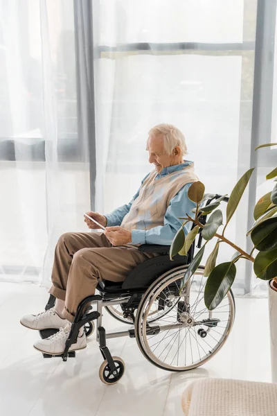 Homem sorridente sênior sentado em cadeira de rodas e usando tablet digital em casa de repouso — Fotografia de Stock