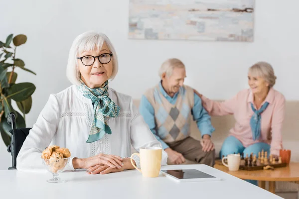 Пожилая женщина в очках пьет чай с печеньем, пока пожилая пара играет в шахматы — стоковое фото