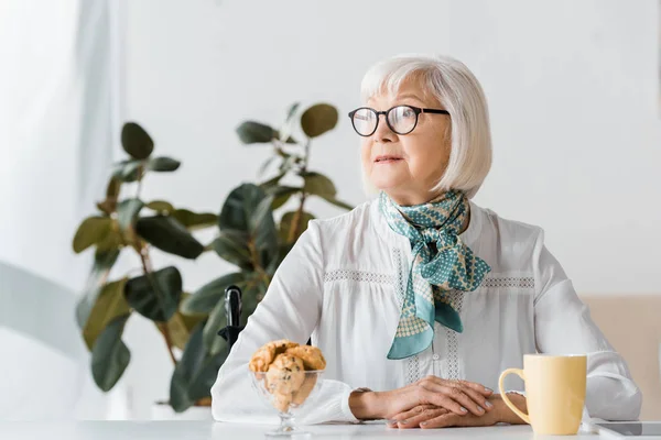 Пожилая женщина в очках сидит за столом с чашкой и печеньем — стоковое фото