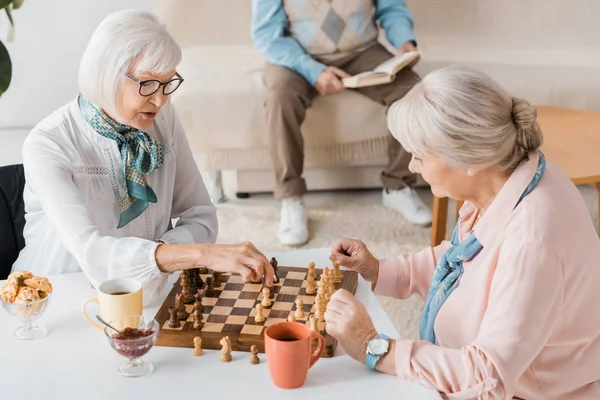 Seniors jouant aux échecs et buvant du café tandis que l'homme lisant le livre sur le canapé — Photo de stock