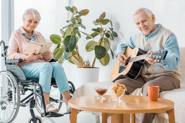 Старша жінка сидить у інвалідному кріслі і читає книгу, а старший чоловік грає на акустичній гітарі — стокове фото