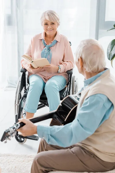 Seniorin sitzt im Rollstuhl und liest Buch, während Senior Akustikgitarre spielt — Stockfoto