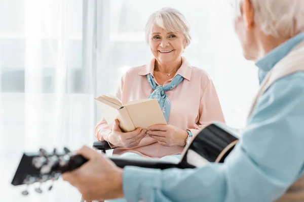 Lächelnde Seniorin mit Buch hört Senioren zu, die Akustikgitarre spielen — Stockfoto