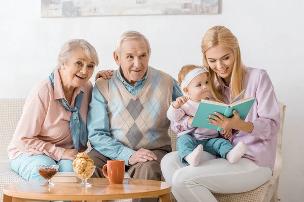 Happy family sitting on sofa and reading book with toddler — Stock Photo