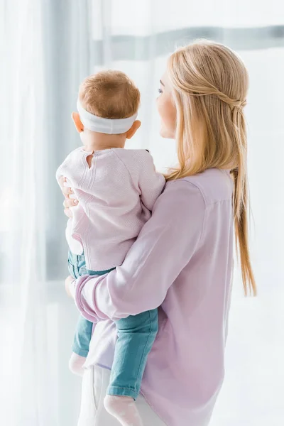 Giovane madre che tiene in braccio il bambino e guarda la finestra — Foto stock