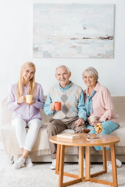 Mujer joven sentada en el sofá y bebiendo té con padres mayores - foto de stock