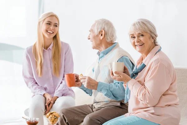 Young woman talking and drinking tea with senior parents on sofa — Stock Photo